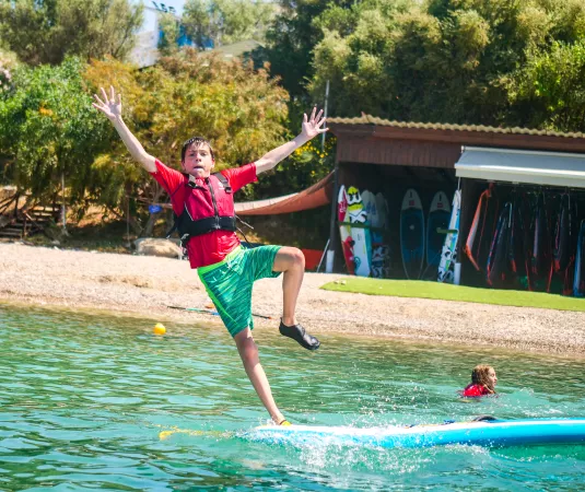  a young boy wearing a red shirt, green shorts, and a life jacket, slipping or falling off a blue paddleboard into the water.