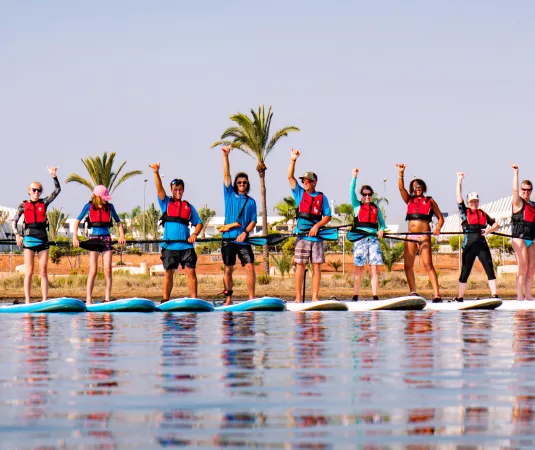 A group of 14 people standing on paddleboards lined up side by side on calm water.