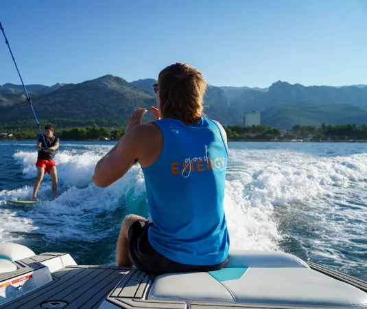 two people engaged in a watersport activity on a sunny day. One person, wearing a blue sleeveless shirt, is seated on the back of a boat, is instructing the other, who is wakeboarding on the water