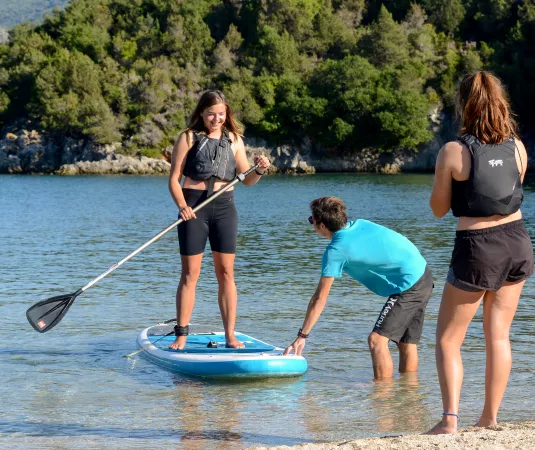 a young girl standing on a blue paddleboard in shallow water near the shoreline, holding a paddle and smiling. A man, bent over, appears to be stabilizing or adjusting the paddleboard in the water.