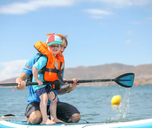 A child and an adult paddleboarding on calm water under a clear blue sky. 