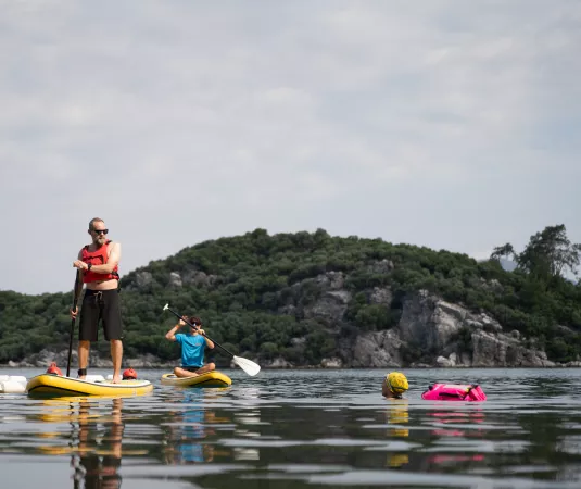 shows two people engaging in paddleboarding on a calm body of water near green, rocky hills under a cloudy sky. One person, standing on a yellow paddleboard, is holding a paddle and wearing sunglasses, a red life jacket, and dark shorts.
