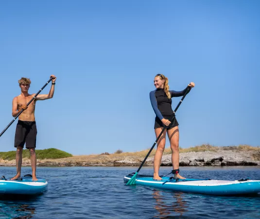 two young individuals paddleboarding on calm blue water under a clear sky. 