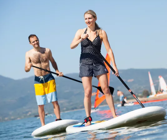 A woman and a man paddleboarding on calm water near a beach.