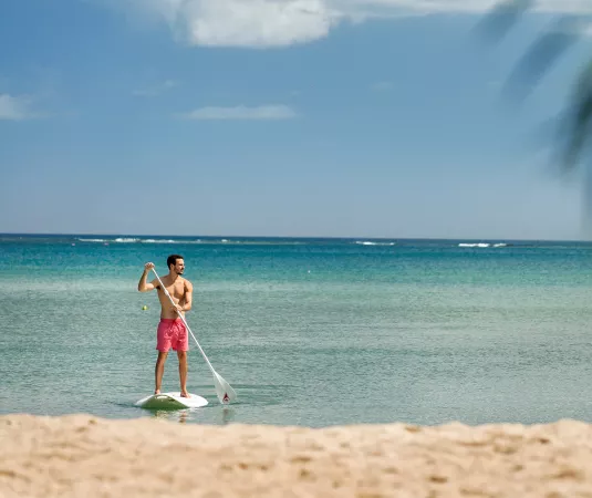  a man paddleboarding on calm, clear turquoise waters near a sandy beach. 