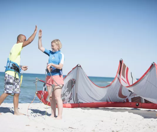 The image shows two people on a sandy beach next to a large, partially deflated red and white kiteboarding kite. A man in a yellow shirt and checkered shorts is giving a high-five to a woman wearing a blue and white top and light pink shorts. Both individuals are barefoot and appear to be preparing for kiteboarding. 