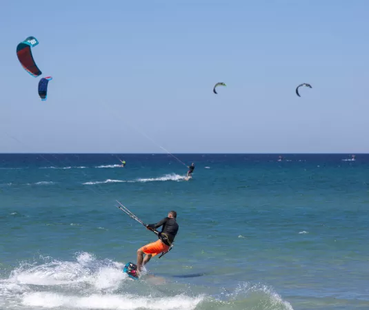 The image shows a vibrant scene of kiteboarding on a sunny, clear day at the sea. In the foreground, a kiteboarder wearing bright orange shorts and a black wetsuit is skillfully riding the waves, holding onto the kite's control bar