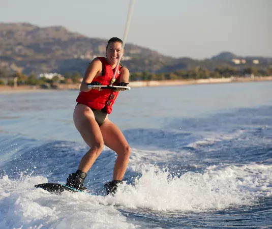 a woman wakeboarding on a body of water near a shoreline with hills and scattered buildings in the background. 