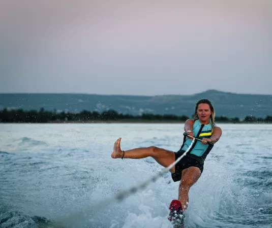  person water skiing on a calm body of water during what appears to be early morning or late evening, as the sky has a soft gradient from gray to light blue. The skier, wearing a life jacket, is holding onto a tow rope attached to a boat that is out of frame. They are balanced on one water ski, leaning back slightly with legs extended for stability. 