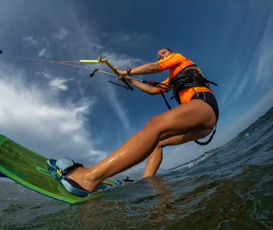 A woman kitesurfing on the ocean, the kite is high in the sky and the photo is taken from a low angle near the water, showing the board clearly