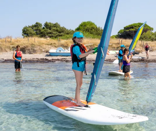 a group of people engaged in windsurfing activities in shallow, clear coastal waters near a rocky shore with dry grass and trees in the background.