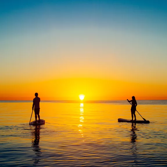 The image captures two people paddleboarding on calm water during a vibrant sunset. 